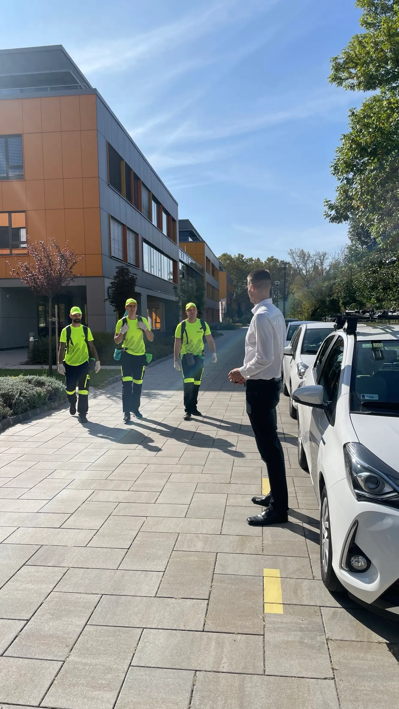 Three workers in bright yellow uniforms approach a man in a white shirt near parked cars outside a modern office building.