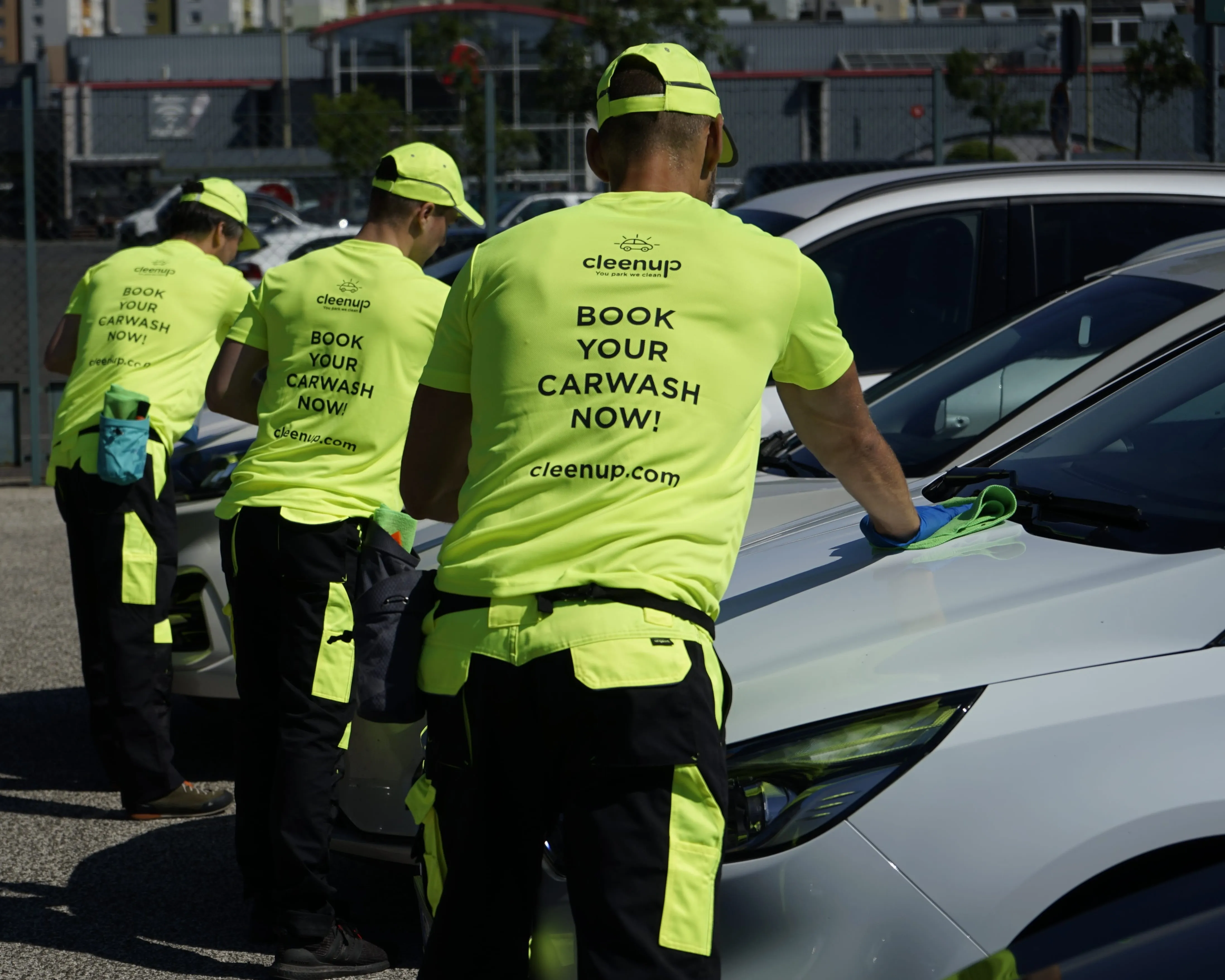 Three workers in bright yellow uniforms clean a white car in an outdoor parking lot.
