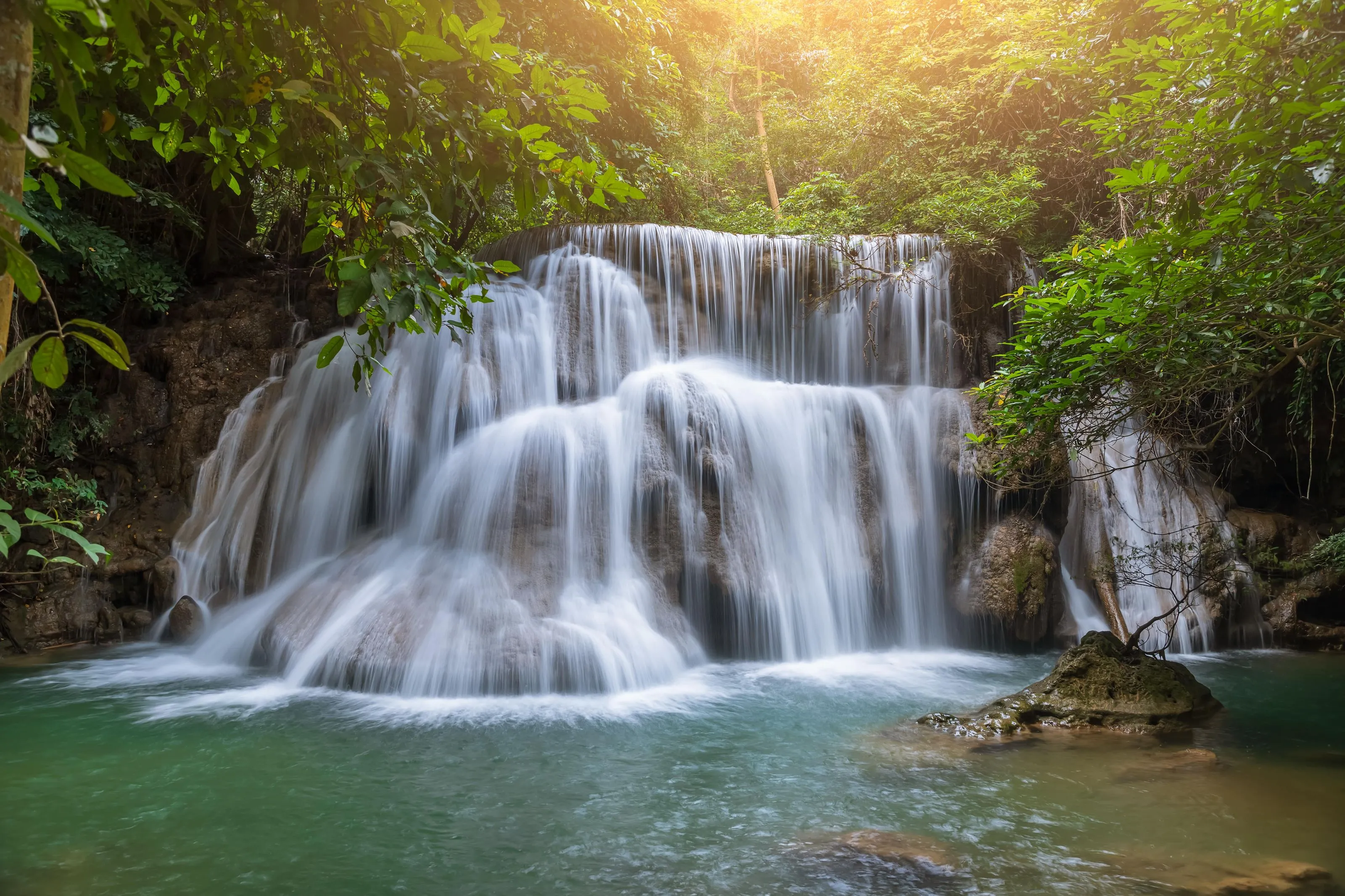 A serene multi-tiered waterfall cascades through lush green jungle foliage, illuminated by golden sunlight filtering through the canopy.